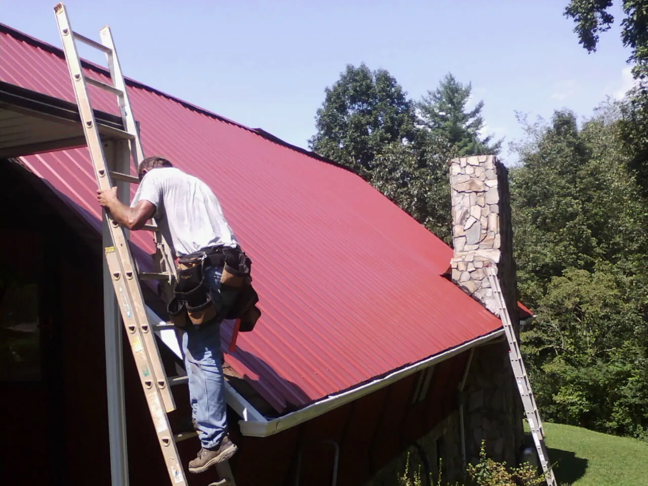 Roofer on a ladder beside a red metal roof and stone chimney — Soffit & Fascia Repair by Roofing Brothers in Mount Angel
